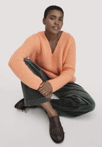 Frau mit short hair sitzt im Schneidersitz, trägt einen orangefarbenen strukturierten Pullover, dunkelgrüne Hosen und dunkelbraune Fransenloafers vor weißem Hintergrund.