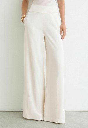 Femme portant un pantalon blanc taille haute à jambes larges, debout contre un mur blanc uni, mains dans les poches.