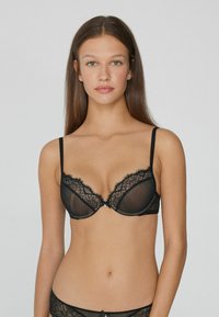 Sostén de encaje negro con diseño a rayas, con copas transparentes, tirantes ajustables y un pequeño lazo decorativo en el centro.