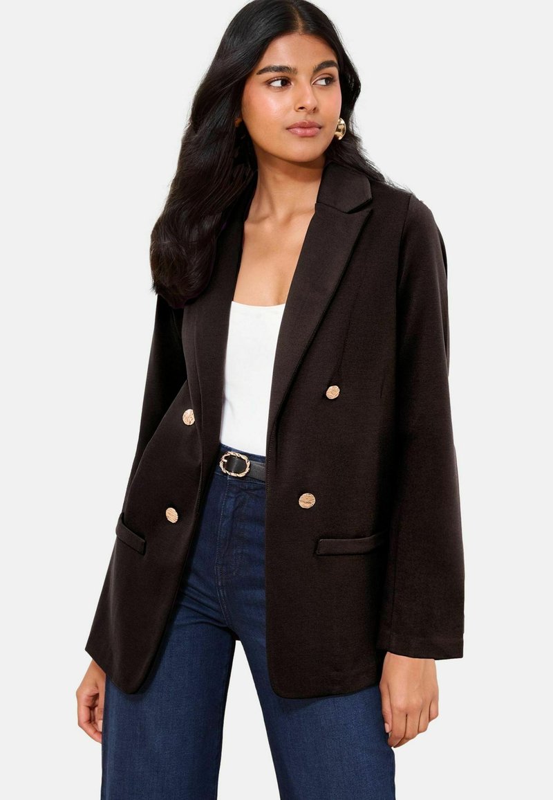 Schwarzer doppelt geknöpfter Blazer mit Revers, goldenen Knöpfen und Fronttaschen, kombiniert mit einem weißen Oberteil und dunklen Jeans.