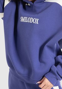 Purple hooded sweatshirt with white detailing, features "SMILODOX" embroidered on the chest, made from a soft, textured fabric.
