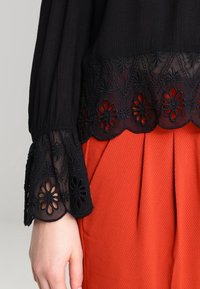 Svart topp med intrikat blommig broderi på ärmarna, kombinerad med en texturerad orange kjol. Designen har en scallopad fåll.