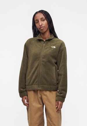 Donna con capelli intrecciati che indossa una giacca in pile verde oliva con zip e pantaloni marrone chiaro, in piedi davanti a uno sfondo semplice.