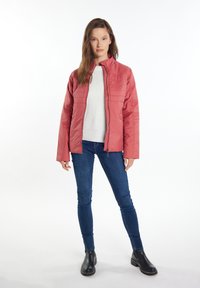 Jachetă ușoară, matlasată, de culoare corai, cu guler înalt și fermoar complet, purtată cu un pulover crem și blugi albaștri. Textură elegantă și fină.
