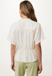 Blusa beige chiaro a maniche corte con vita arricciata e pieghe posteriori, realizzata in un tessuto liscio. Indossata sopra pantaloni verde chiaro.