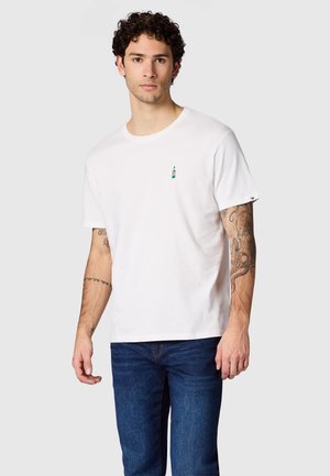 Jonge man met krullend haar draagt een wit T-shirt met een klein groen fleslogo en blauwe jeans, staand tegen een grijze achtergrond.