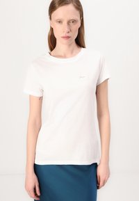 T-shirt bianco in cotone con scollo a girocollo e maniche corte, caratterizzato da un piccolo logo grigio chiaro sul petto. Texture liscia.