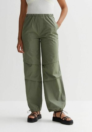 Žena má na sobě olivově zelené kalhoty širokého střihu s elastickými manžetami a černé sandály na platformě, stojí s jednou rukou v kapse.