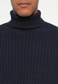 Pull ras du cou marine en maille texturée avec un design côtelé, présentant un col ajusté et une forme cylindrique.