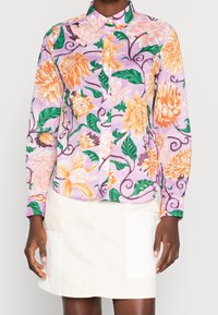 Chemise à boutons à motif floral avec une base lilas, des fleurs orange et roses, des feuilles vertes et un col. Portée avec une jupe légère.