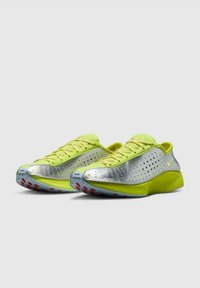 Un paio di scarpe sportive neon gialle e argento con lati perforati, lacci e accenti rossi sulla suola traslucida, angolate a sinistra su una superficie grigia.