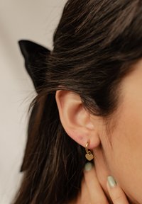 Orecchino a cerchio in oro con un piccolo ciondolo a forma di cuore, indossato sul lobo. I capelli sono scuri e coprono parzialmente l'orecchio. Le unghie sono verniciate di verde.