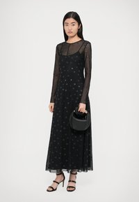 Čierne maxi šaty s bielymi bodkami, s dlhými priehľadnými rukávmi, priliehavým živôtikom a splývavou sukňou. Doplnky zahŕňajú malú čiernu kabelku a sandále na remienky.