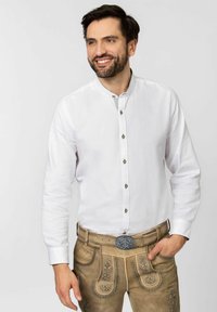 Weißes langärmliges Hemd mit Stehkragen, versehen mit dekorativen Knöpfen. Getragen mit beigen Lederhosen, die Steppnähte und Akzente zeigen.