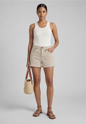 Vrouw draagt een wit hemdje, beige versleten korte broek, bruine sandalen, houdt een geweven mandtas met leren riempjes vast, staat op een grijze achtergrond.