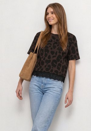 Jeune femme aux longs cheveux bruns portant un haut noir et marron à motifs, un jean bleu clair et un sac à bandoulière marron clair.