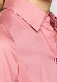 Camicia rosa chiaro con una texture liscia, colletto classico e bottoni. Presenta un design aderente e un leggero lucido.