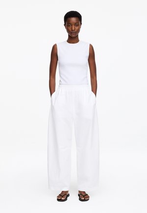 Femme portant un top blanc sans manches et un pantalon blanc à jambes larges, les mains dans les poches, debout sur un fond blanc.