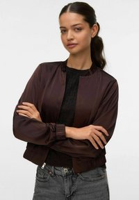 Giubbotto bomber satin marrone con colletto e polsini a costine, dotato di chiusura con zip. Indossato sopra un maglione scuro a trama e jeans grigi.