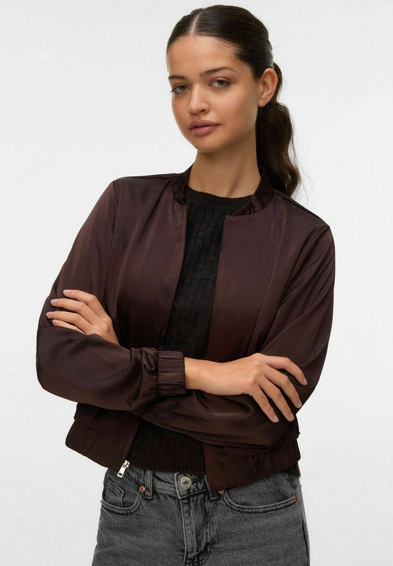 Giubbotto bomber satin marrone con colletto e polsini a costine, dotato di chiusura con zip. Indossato sopra un maglione scuro a trama e jeans grigi.