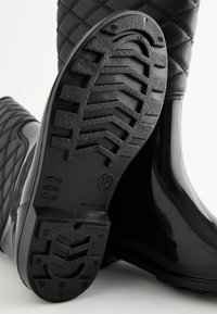 Des bottes en caoutchouc noires et étanches présentent une texture supérieure matelassée, un bout rond et une semelle texturée à motifs en relief pour une meilleure adhérence.