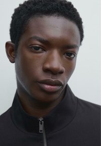 Black zip-up jacket with a high collar, featuring smooth texture and silver zipper. Close-up of a face with neutral expression and short, curly hair.