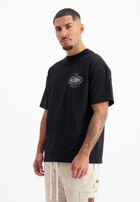 Zwart katoenen T-shirt met een ronde hals, korte mouwen en een decoratieve logo in paars en goud aan de voorkant links.