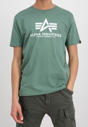 Mężczyzna w zielonej koszulce Alpha Industries z białym logo i ciemnych spodniach typu cargo, jedna ręka w kieszeni, na jednolitym tle.