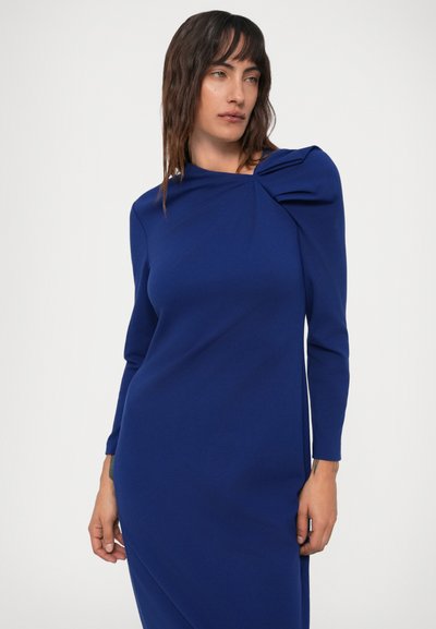 Mulher com cabelo escuro de comprimento médio, vestindo um vestido justo de mangas longas na cor azul-royal, com um detalhe franzido no ombro esquerdo.