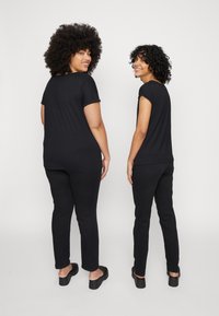 Dois modelos vestem camisetas pretas de manga curta e calças pretas, exibindo uma textura suave e um corte descontraído. Ambos os modelos usam tamancos pretos.