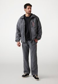 Jackete gris con capucha y puños elásticos, combinado con pantalones gris a juego. Incluye un acento naranja y detalles del logo. Llevado con zapatillas negras.