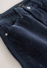 Pantalon en velours côtelé bleu foncé présentant un design classique à cinq poches, avec des coutures ton sur ton et des détails en métal sur la ceinture.