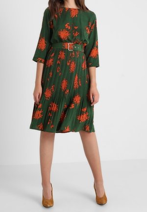 Frau trägt ein grünes Midikleid mit orangefarbenem Blumenmuster, plissiertem Rock, dreiviertellangen Ärmeln, tailliertem Gürtel und braunen Absätzen.