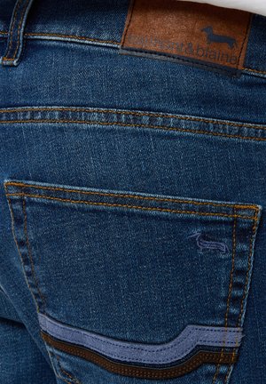 Jeans de mezclilla de color azul oscuro, con costuras contrastantes, un parche de cuero con un logo en relieve y un bolsillo con un ribete decorativo.