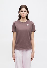 Νεαρός άνθρωπος που στέκεται με το πρόσωπο προς το κοινό, φορώντας ένα μοβ μπλουζάκι Nike και ανοιχτό ροζ παντελόνι, μπροστά από ένα απλό λευκό φόντο.