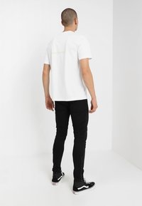 Camiseta blanca de algodón con un pequeño gráfico de texto verde en la parte posterior, combinada con unos jeans negros ajustados y zapatillas negras.