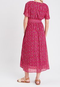Femme portant une robe midi à motifs roses avec des manches courtes et des sandales marron, se tenant de dos sur un fond blanc.