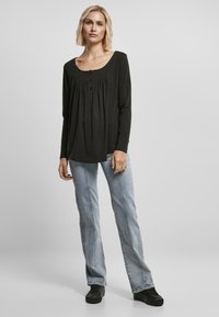 Zwarte blouse met lange mouwen en een ronde hals, met plooien en een knoopdetail, gecombineerd met lichtblauwe jeans en zwarte schoenen.