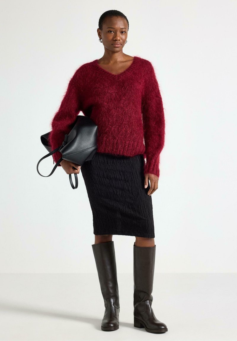 Viininpunainen, pörröinen neule V-pääntiellä, yhdistetty mustaan, teksturoituun kynähameeseen ja polven korkeisiin ruskeisiin nahkasaappaisiin. Musta käsilaukku kädessä.