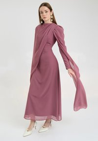 Długa, zwiewna sukienka z miękkiego, drapowanego materiału w kolorze mauve, z wysokim dekoltem, bufkami i warstwami przezroczystych akcentów. Połączona z białymi szpilkami.