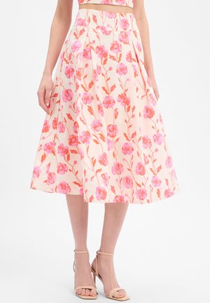 Vrouw draagt een hoog-tailleroze, geplooide midi-rok met roze en oranje bloemenprint en nude enkellaarzen met hoge hak tegen een witte achtergrond.