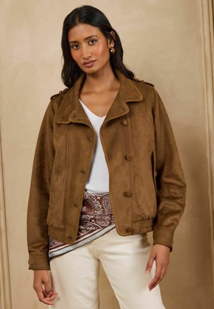 Woman with long dark hair wears brown buttoned jacket over white top and paisley scarf, paired with light beige pants, standing indoors.