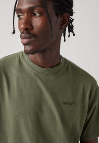 Tricou din bumbac de culoare verde măsliniu, cu guler rotund, având un logo Levi's subtil imprimat în aceeași culoare pe piept. Textură netedă.