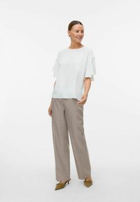 Haut blanc texturé à manches courtes à volants, associé à un pantalon large marron clair et des ballerines vertes, se tenant devant un fond uni.