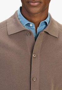 Bruine geribde cardigan met een kraaghalslijn, met vijf knopen, gedragen over een lichtblauw overhemd met kraag. Glad textuur.