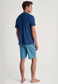 Marineblauwe katoenen t-shirt gecombineerd met gestreepte blauwe shorts met een geometrisch patroon. Model staat naakt met een neutrale achtergrond.