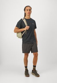 Camisa de manga corta gris oscuro con un logo, combinada con pantalones cortos oscuros. Mochila de color claro con tirantes sobre un hombro y zapatillas deportivas grises.