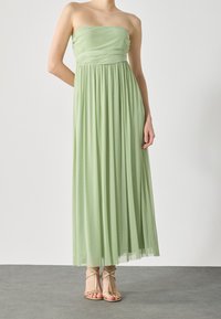 Femme portant une robe longue fluide vert clair sans bretelles avec des plis, assortie à des sandales dorées à talons hauts et à lanières.