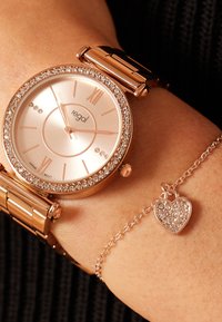 Roze gouden polshorloge met een kristallen bezel, ronde wijzerplaat en Romeinse cijfers. Vergezeld van een delicate armband met een hartbedel.