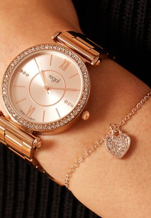 Rose gold wristwatch with a crystal bezel, circular face, and Roman numerals. Accompanied by a delicate bracelet with a heart charm.